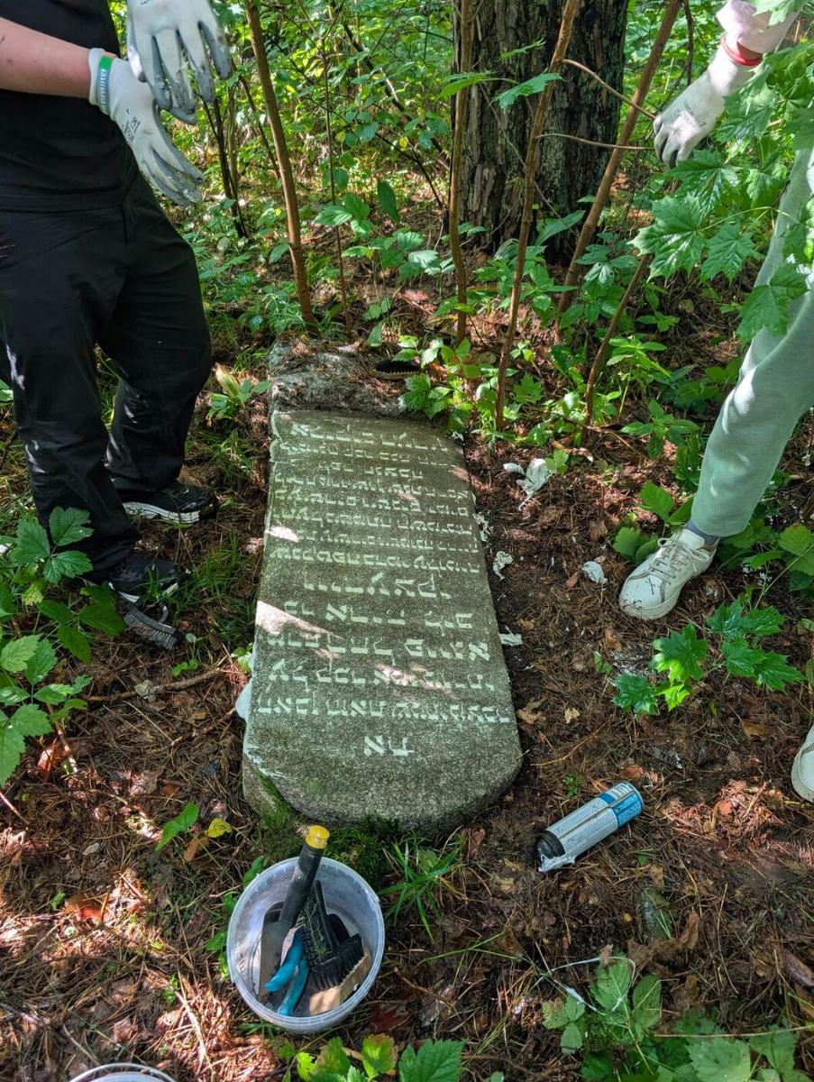 A cleaned slab in the forest with its Hebrew inscription legible again, brushes in a bucket beside it