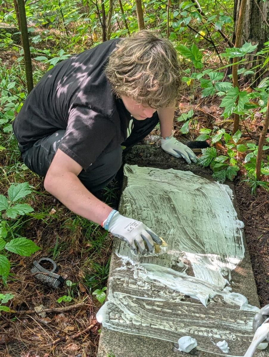 Jules kneeling in the forest, brushing cleaning solution across a fallen matzevah
